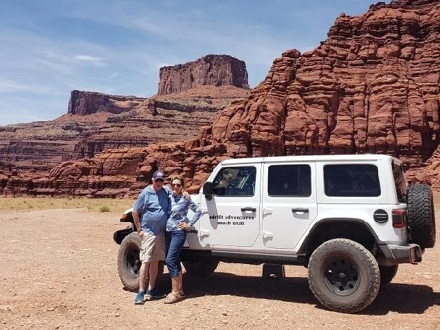 Ein Paar posiert neben einem weißen Jeep mit roten Felsklippen im Hintergrund
