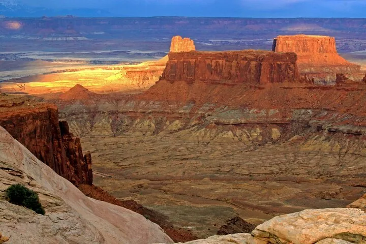 Sunlight illuminates a prominent red rock mesa in a vast canyon landscape